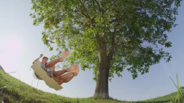 Slow Motion close-up lage hoek: lachende vriendin en vriend op een date wuivende onder een grote oude boom. Grinning jong stel genieten van verse en lente lucht groene natuur op houten touw Swing. - Video