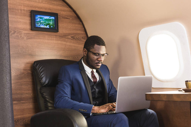 Attractive and successful African American businessman with glasses working on a laptop while sitting in the chair of his private jet. - Foto, Bild