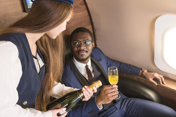 Молодий успішний афро-американський бізнесмен в окулярах і стюардеса показує пляшку вина в салоні приватного струменя. Обслуговування і політ першого класу. - Фото, зображення