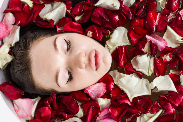 A young attractive girl takes a bathroom with flower petals and relaxes against the background of a beautiful light interior. Spa treatments for beauty and health with skin care - Foto, Imagen