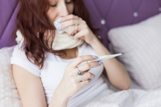 An attractive young brunette with brown hair sits in her bed with a thermometer. Colds and home treatment - Photo, Image