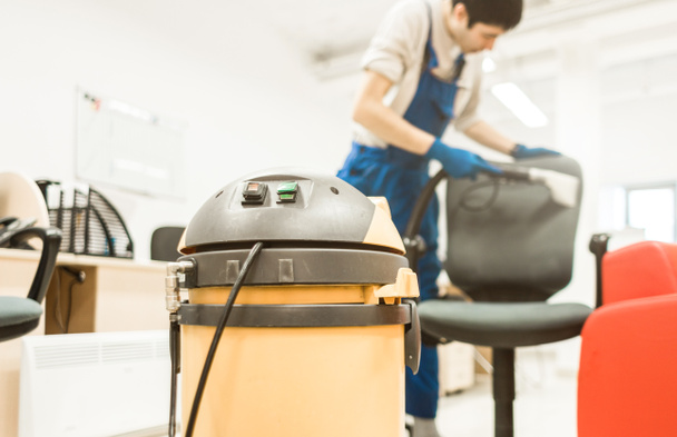 Young man in workwear and rubber gloves cleans the office chair with professional equipment. - 写真・画像