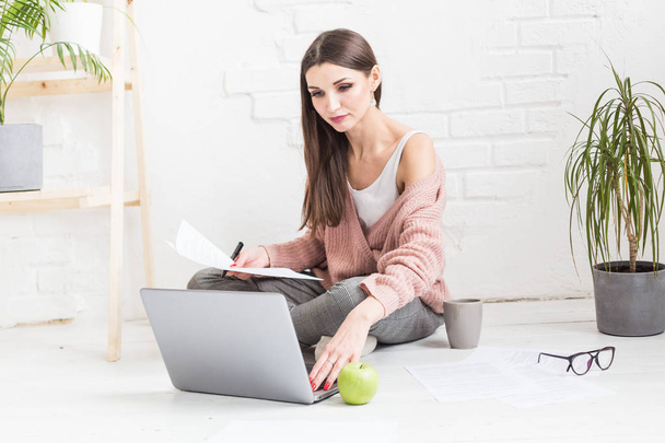 A young happy woman sits on the floor in a bright apartment or office interior and works at a laptop, freelancer girl at work, the concept of distance learning students, paperwork and online learning - Foto, Imagen
