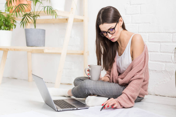 A young happy woman sits on the floor in a bright apartment or office interior and works at a laptop, freelancer girl at work, the concept of distance learning students, paperwork and online learning - Фото, зображення