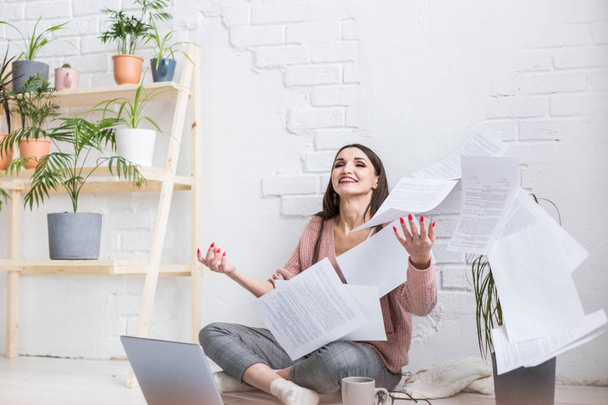 A young happy woman throws up a bunch of pieces of paper and documents sitting near the laptop, the freelance girl is happy and in a good mood. excitement and inspiration - Foto, Bild