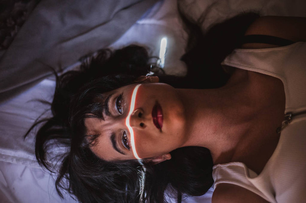 jonge vrouw in bed met een lichtbundel over haar gezicht en intense blik - Foto, afbeelding