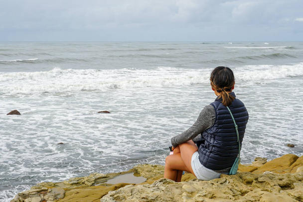 Молода азіатка сиділа на скелі і дивилася далеко на море. Красива дівчина дивиться з моря на пляж. надихаючий момент. Портрет молодої жінки, що дивиться на горизонт. - Фото, зображення