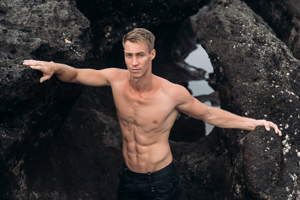 Handsome muscular man with bare-chested posing near rock on black sandy beach - Φωτογραφία, εικόνα
