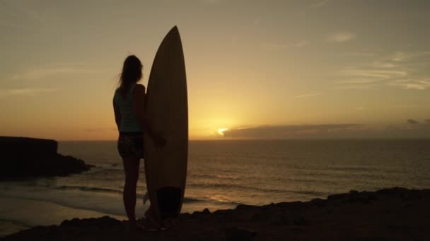 Yavaş Hareket: Sörfçü kız büyüleyici görünümüne bakan bir okyanus uçurumun tepesinde duran bir sörf tahtası tutan. Dalgalar sürme uzun bir günün ardından zihnini temizleyen bir güverte ile genç kadın atlet. - Video, Çekim