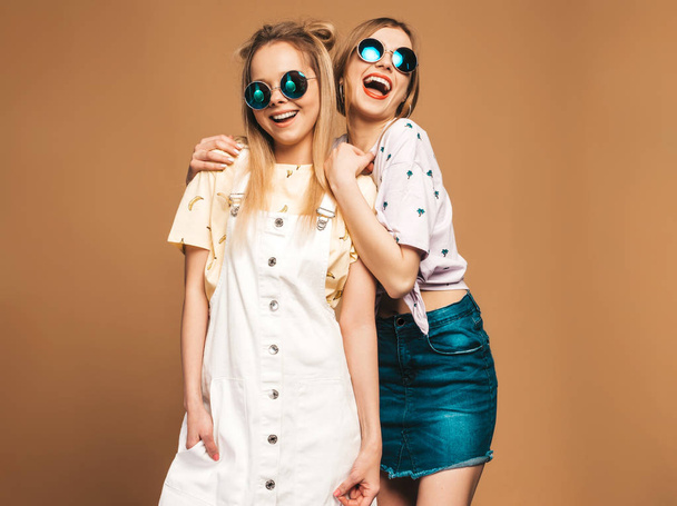 Two young beautiful smiling blond hipster girls in trendy summer colorful T-shirt clothes. Sexy carefree women posing near beige wall in round sunglasses. Positive models having fun - Photo, Image