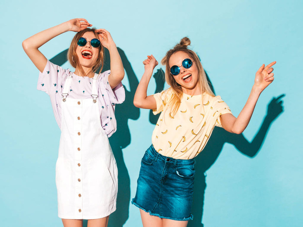 Two young beautiful smiling blond hipster girls in trendy summer colorful T-shirt clothes. Sexy carefree women dancing near blue wall in round sunglasses. Positive models having fun - Foto, Imagem