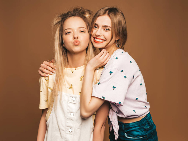 Two young beautiful smiling blond hipster girls in trendy summer colorful T-shirt clothes. Sexy carefree women posing on beige background. Positive models giving kiss - Foto, Bild