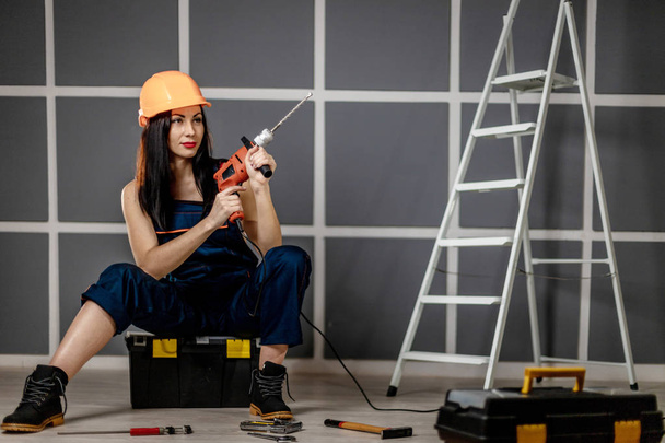 Beautiful brunette women, dressed in construction uniform and orange helmet with drill and tools box working in office room - Photo, Image
