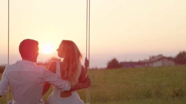SLOW MOTION CLOSE UP: Joyous girlfriend and boyfriend kissing and laughing on swing on summer evening in countryside. Young couple enjoying last sunrays on romantic date whilst swaying on rope swing. - Πλάνα, βίντεο