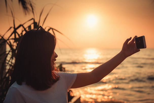Retrato de mulher está usando Smartphone e selfie-se na praia durante o pôr do sol, turista asiático está relaxando com seu telefone celular na praia em tempo de férias. Conceito de comunicação tecnológica
. - Foto, Imagem
