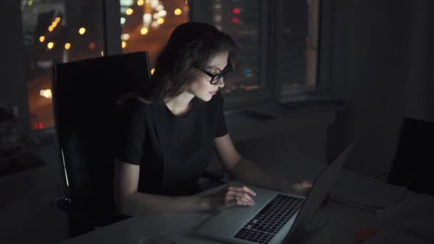 Porträt eines jungen Mädchens, das spät im Büro arbeitet. Geschäftsfrau mit Brille arbeitet an einem Laptop vor dem Hintergrund der nächtlichen Stadt - Filmmaterial, Video