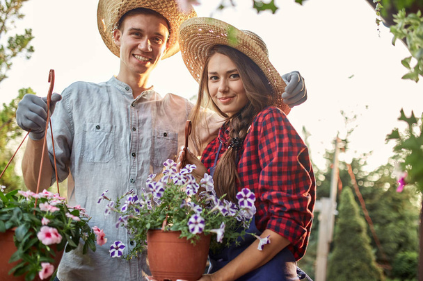 Guy and girl gardeners in a straw hats hold pots with wonderful petunia on the garden path in  on a sunny day. - Foto, Imagem