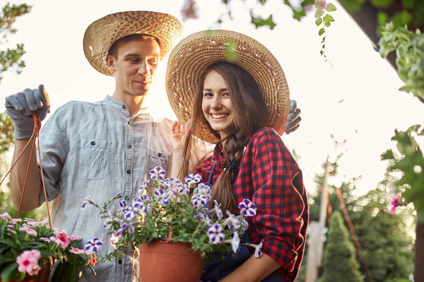 Guy and girl gardeners in a straw hats hold pots with wonderful petunia on the garden path in  on a sunny day. - Fotó, kép