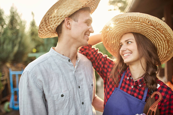 Happy guy and girl gardeners in a straw hats look at each other in the garden on a sunny day. - Foto, afbeelding