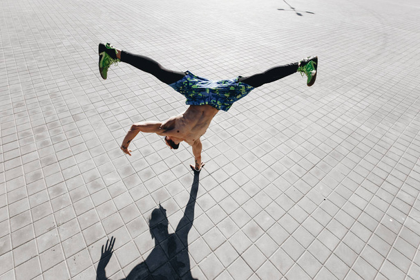 Young athletic man with a naked torso with tattoos dressed in the  black leggings and blue shorts doing the trick standing on one hand on the paving slabs in the street - 写真・画像