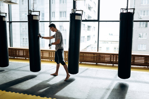 Guy dressed in the grey t-shirt and black shorts stands on guard and works out a boxing punch next to hanging punching bag against the background of panoramic windows in the gym - Foto, afbeelding
