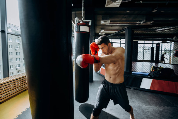 Guy in red boxing gloves with a naked torso dressed in the black shorts hits punching bag  in the gym  against the background of panoramic windows - Foto, Bild