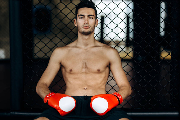 Sportsman with a naked torso and the red boxing gloves  on his hands  sits on the border of boxing ring - Φωτογραφία, εικόνα