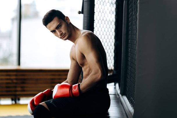 Brutal dark-haired guy with a naked torso and the red boxing gloves  on his hands  sits on the border of boxing ring - Фото, изображение