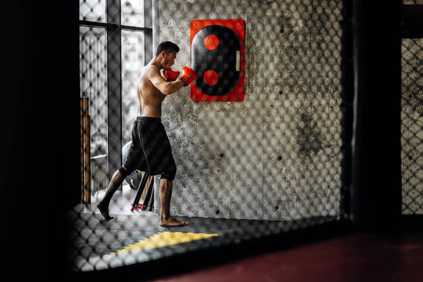 Sportsman with a naked torso and in the red boxing gloves hits equipment for boxing on a concrete wall in the gym behind the net - Фото, изображение