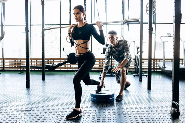 Slim dark-haired girl dressed in black sportswear is doing barbell squat and the coach helps her in the gym - Fotoğraf, Görsel