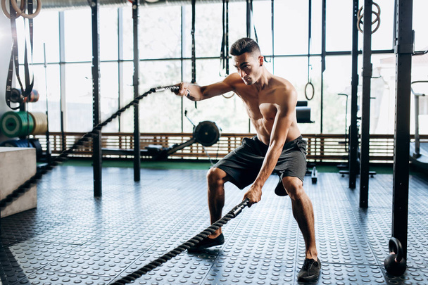 Jovem atlético com um tronco nu vestido com shorts pretos fazendo exercícios esportivos com cordas esportivas no ginásio
 - Foto, Imagem