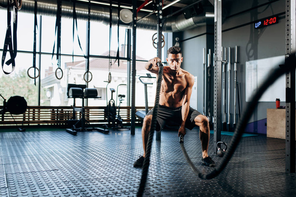 Athletic dark-haired man with a naked torso dressed in the  black shorts doing sport exercises with sports ropes in the gym - Foto, Bild