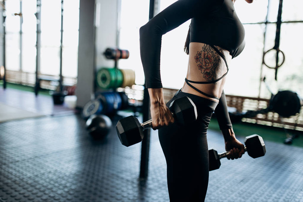 Slender body of a young woman with tattoo in a black sportswear that is doing exercises with dumbbells in the gym - Valokuva, kuva