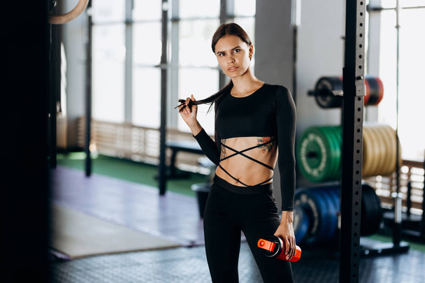 Athletic dark-haired girl dressed in black sportswear stands with water in her hand near the sport equipment in the gym and touches hair - Foto, immagini