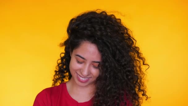 Portrait of young latin girl with curls flirts on yellow background. Tempting woman smiling to camera. Studio footage. Female in red t-shirt, casual style. - Кадры, видео