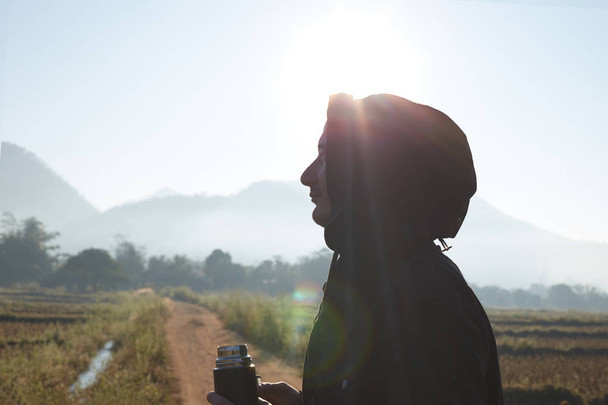 Προφίλ close up πορτρέτο του αρσενικού ταξιδιώτη ντυμένος με μαύρο μπουφάν με κουκούλα, κρατώντας θερμό μπουκάλι με ζεστό τσάι, κοιτάζοντας γύρω, αναπνέει φρέσκο πρωινό αέρα στα βουνά, απολαμβάνοντας τη φύση μόνο. - Φωτογραφία, εικόνα