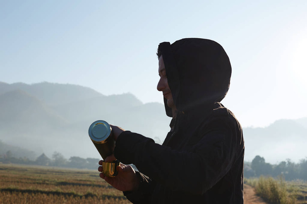 Perfil close up retrato de homem viajante vestido com jaqueta preta com capuz derramando bebida quente de copo termo, respirando ar fresco da manhã nas montanhas, desfrutando da natureza sozinho, caminhadas ou trekking
.  - Foto, Imagem