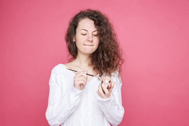 Attractive curly haired young female paints traditional Easter egg with brush brush, preparing gifts for coming holiday, wearing cozy knitted white outfit. Easter and spring celebration concept. - Photo, Image