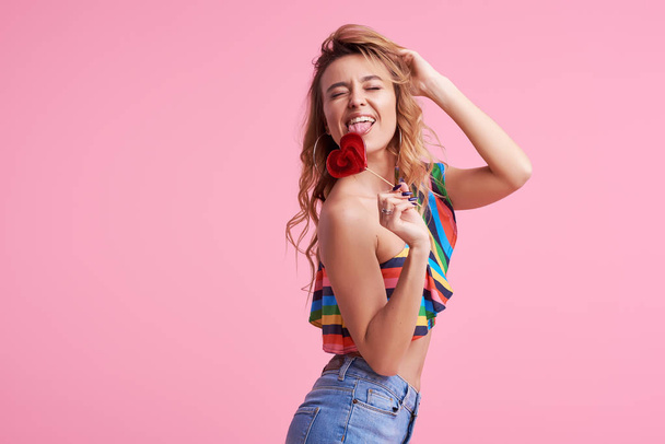 Sweet and candy. The girl colorful dress on pink background in the studio. Woman holding a red heart-shaped lollipop. Valentine's Day. Girl holding a lollipop in open mouth. Wow and shocked concept.  - Photo, Image