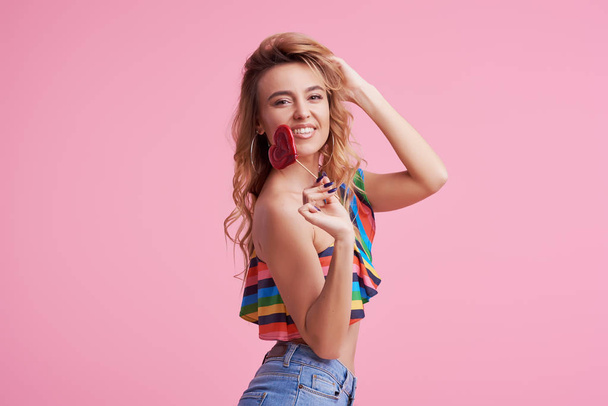 Sweet and candy. The girl colorful dress on pink background in the studio. Woman holding a red heart-shaped lollipop. Valentine's Day. Girl holding a lollipop in open mouth. Wow and shocked concept.  - Foto, Imagem