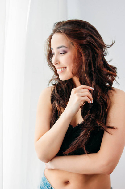 Счастливая чувственная улыбающаяся девушка азиатская молодая женщина с темными длинными вьющимися волосами в черном нижнем белье, смотрящая на окно белый занавес
 - Фото, изображение