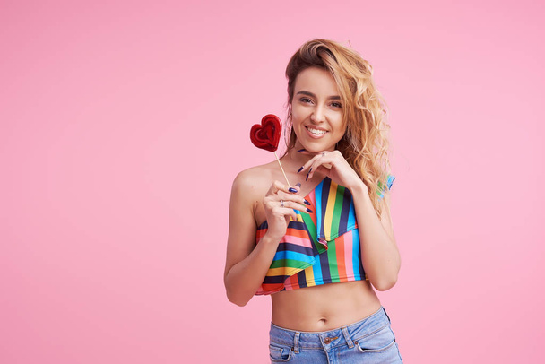 Sweet and candy. The girl colorful dress on pink background in the studio. Woman holding a red heart-shaped lollipop. Valentine's Day. Girl holding a lollipop in open mouth. Wow and shocked concept.  - Foto, immagini
