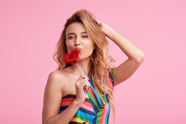 Sweet and candy. The girl colorful dress on pink background in the studio. Woman holding a red heart-shaped lollipop. Valentine's Day. Girl holding a lollipop in open mouth. Wow and shocked concept.  - Fotoğraf, Görsel