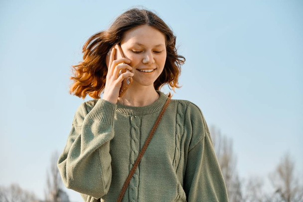 Outdoor portrait of smiling teen girl walking and talking on the phone, blue sky background - Fotó, kép
