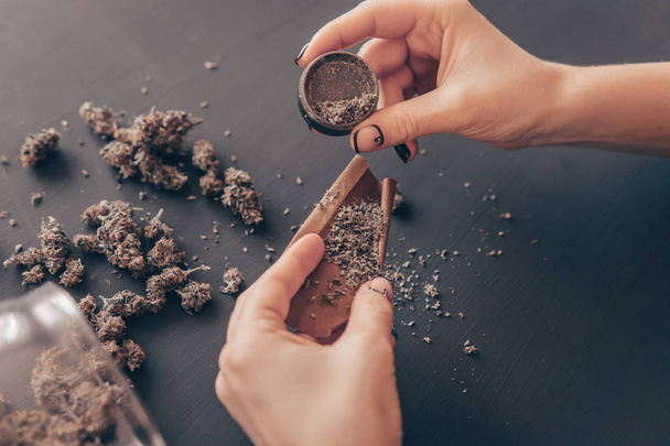 Woman rolling a marijuana joint. Woman preparing and rolling marijuana cannabis joint. Marijuana use concept. Close up of marijuana blunt with grinder. - Фото, изображение