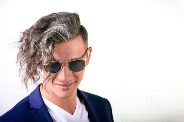 Portrét stylový podnikatel s kudrnaté dlouhé vlasy v sluneční brýle se usmívá na kameru na bílém pozadí - Fotografie, Obrázek