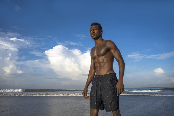 portrait of young attractive and fit black afro American man with strong muscular body posing cool model attitude on the beach isolated on Summer blue sky - Fotó, kép