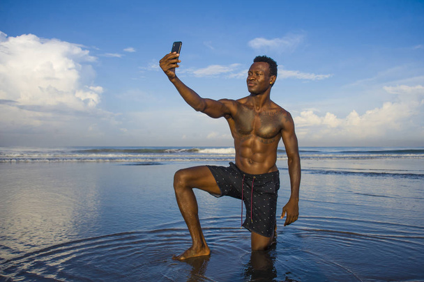 νέοι ελκυστικό και ευτυχισμένο μαύρο αφροαμερικάνικο άθλημα άνθρωπος με αθλητικό σώμα και εξέκτης λήψη φωτογραφία selfie με το κινητό τηλέφωνο στην όμορφη παραλία χαμογελαστή δροσερό στις διακοπές - Φωτογραφία, εικόνα