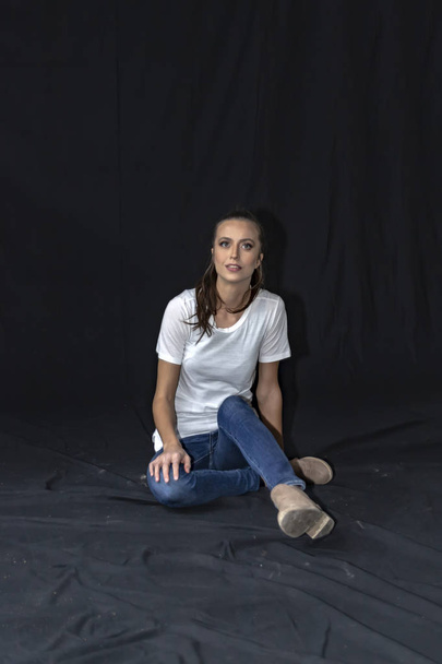Girl in blue jeans and white t-shirt sitting on the floor with crossed legs. - Фото, изображение