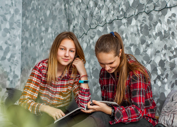 Two Teenage Girls reading fashion Magazine sitting on a couch at home on gray wall background - 写真・画像